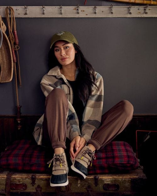 Brunette wearing hiking boots, jobbers, a plaid shacket, tee, and a baseball cap sitting in a rustic indoor home.