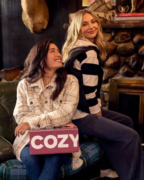 Two women sitting in a cabin wearing cozy outfits from Wantable and holding a Cozytober box.