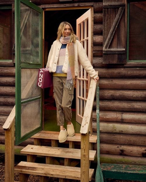 Blonde girl wearing Uggs, a sherpa jacket, cargo pants, a striped sweater, and a scarf from Wantable standing outside a log cabin.