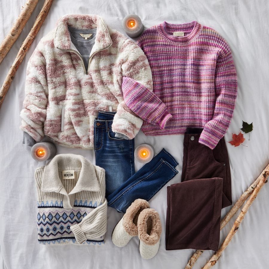 Sherpa quarter-zip pullover, striped sweaters, fair isle sweater, skinny jeans, corduroy pants, and fuzzy slippers laid out in a fall setting with candles and leaves.