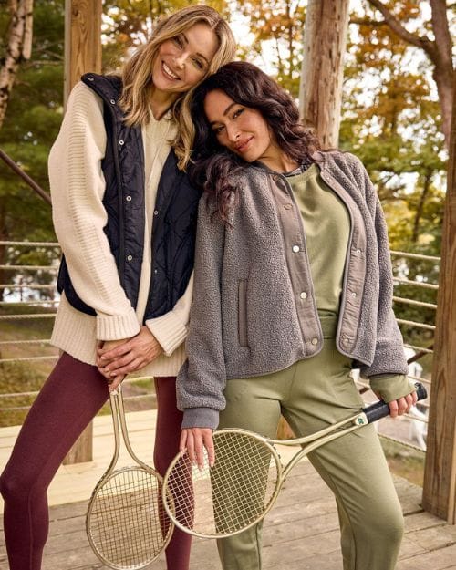 Two happy women wearing cozy activewear holding tennis rackets outside.