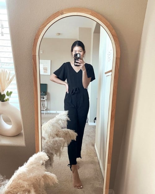 An influencer wearing a versatile black jumpsuit barefoot in her home in front of a mirror. 