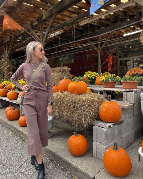 An influencer wearing a matching brown/mauve sweater pants and top set with black boots at a pumpkin patch.