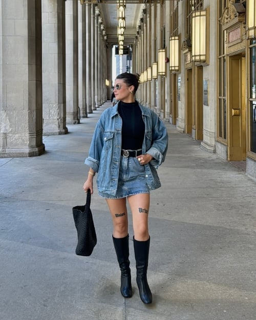 An influencer wearing a denim skirt, oversized denim jacket, a black turtleneck, and black knee boots. 