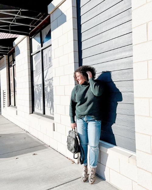 An influencer wearing a green oversized sweater, boyfriend jeans, and snakeskin ankle boots—one of our favorite comfy Thanksgiving outfits.