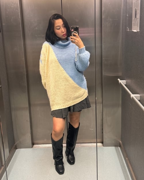 Woman wearing a color-block oversized sweater with a gray skirt and black knee-high boots, taking a mirror selfie.