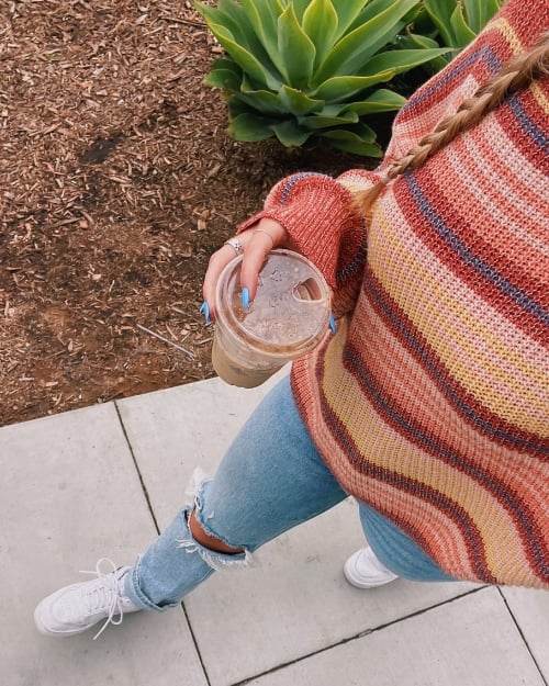 Woman wearing a striped oversized sweater with distressed jeans and white sneakers, holding an iced coffee.