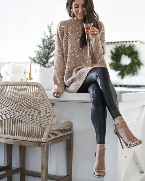 Woman in a sequined oversized sweater and leather leggings, seated on a kitchen counter with a glass of champagne, wearing strappy heels.