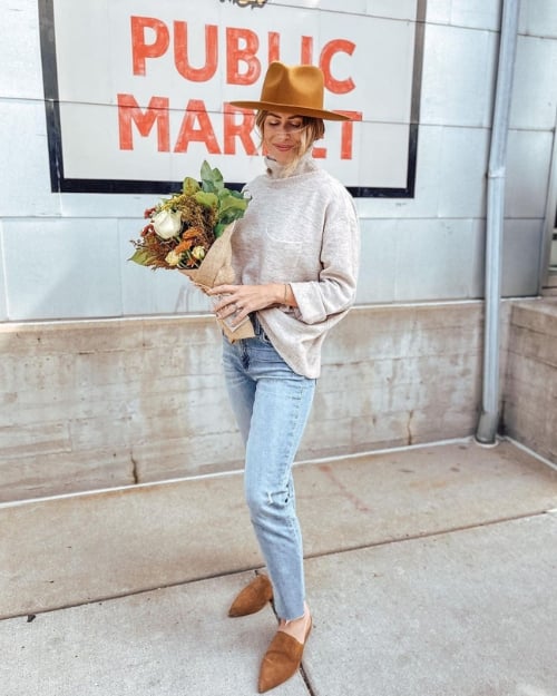 Woman in a tan oversized sweater, light-wash jeans, and a brown hat, holding a bouquet of flowers outside the Milwaukee Public Market.