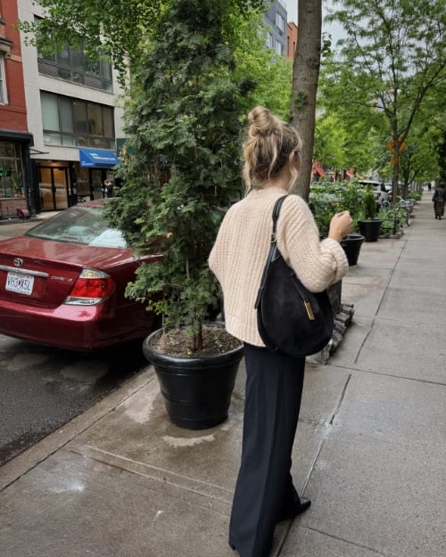 Woman wearing a chunky cream oversized sweater with black wide-leg trousers and a black handbag, walking on a city sidewalk.