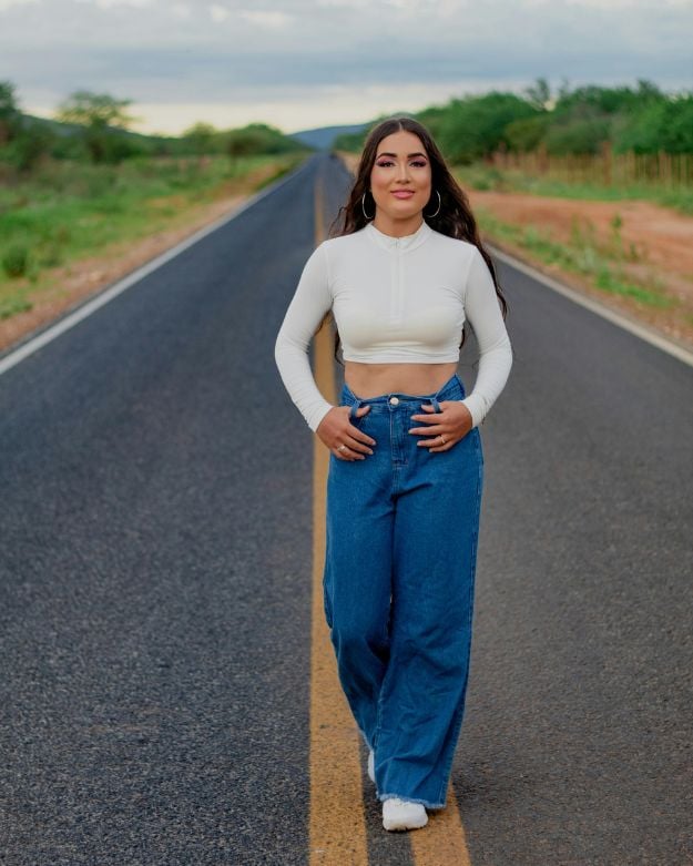 A girl with long brown hair walking down the road wearing a cropped white long-sleeve shirt and wide-leg blue jeans.