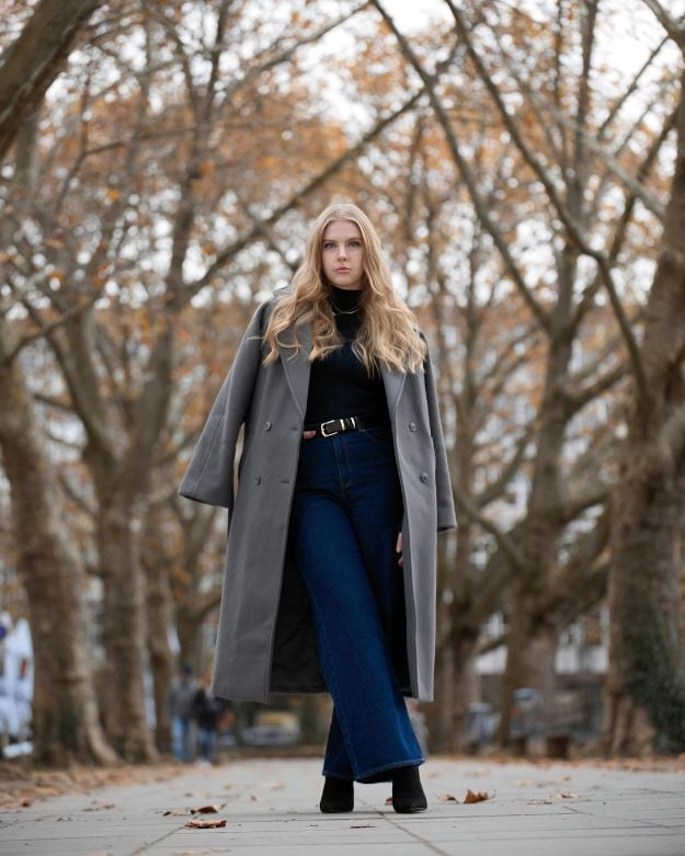 A blonde girl in the forest wearing blue wide-leg jeans, a sweater, and a gray peacoat. 