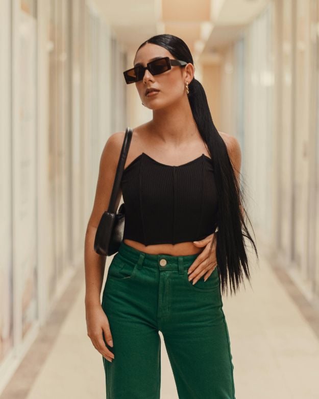 A woman with a long, dark ponytail. and sunglasses wearing a black crop top and forest green denim.