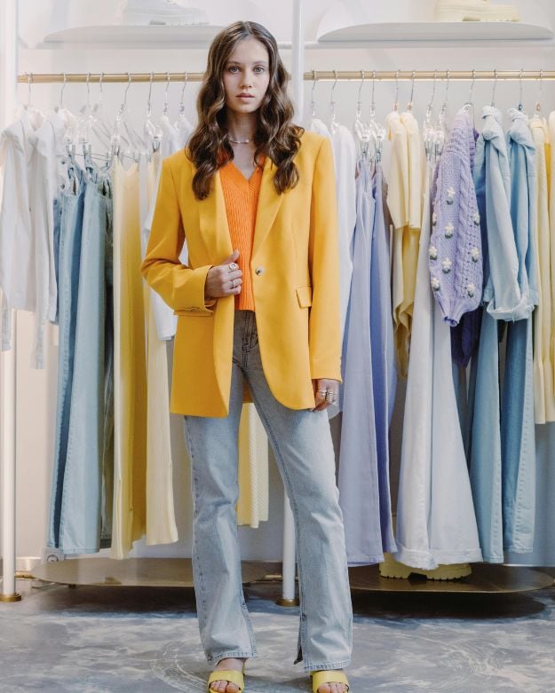 A brown-haired woman wearing blue straight jeans, an orange sweater, and an oversized yellow blazer. 