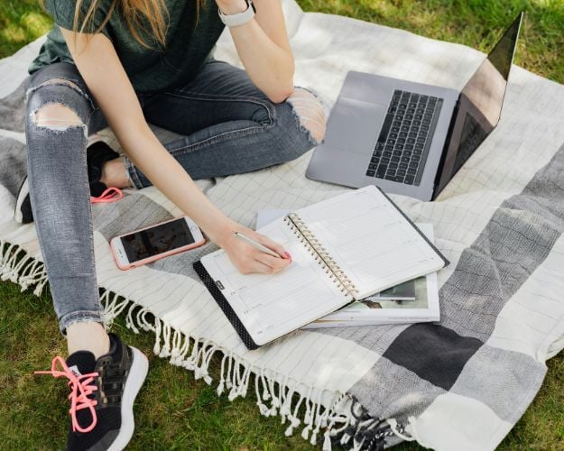 A woman working on her laptop on a blanket in a park wearing gray distressed skinny jeans and sneakers. 