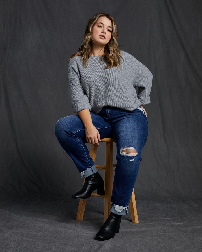 Blonde woman sitting on a stool while wearing dark wash distressed boyfriend jeans and a grey sweater.