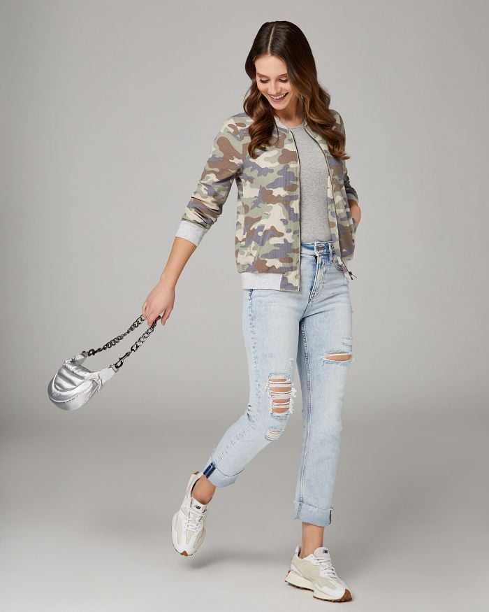 Smiling brunette wearing distressed light wash jeans, grey tank top, and camo bomber jacket while swinging a silver bag.
