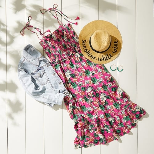 A zoomed-out photo of a light wash denim jacket laying on wooden slats next to a pink ruffled maxi dress with a floral print and a straw hat. 
