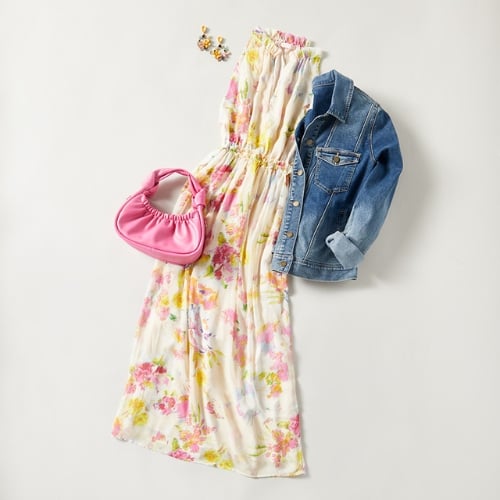 A flat-lay photo showing an example of styling spring dresses. It is a white maxi dress with a floral print next to a medium-wash denim jacket, a pink handbag and a pair of beaded earrings.