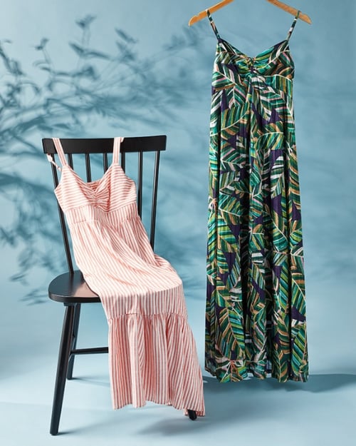 An up-close photo of a striped maxi dress laying across a black chair. Next to it is a tropical print maxi dress hanging from a wooden hanger. 
