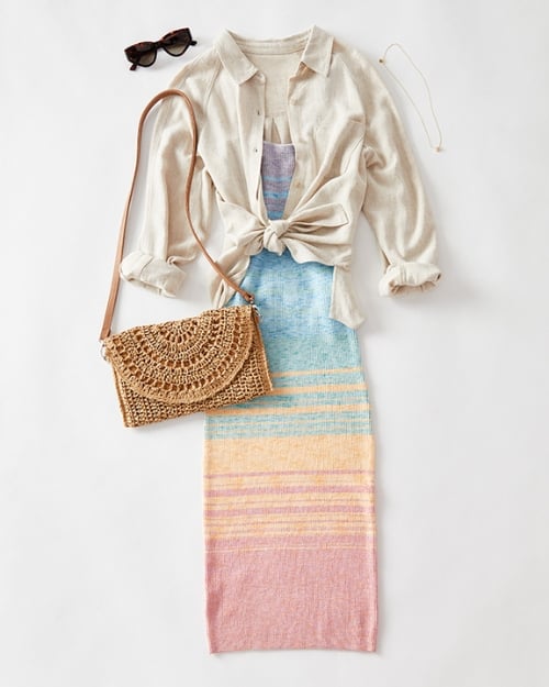 A flat-lay photo showing a bodycon midi dress in pastel colors layered under a white linen long-sleeve shirt. There is also a straw bag and a pair of black sunglasses. Shows how to style spring dresses. 