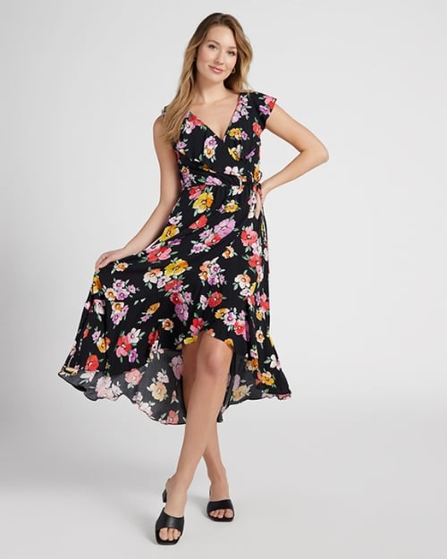 A full-body photo of a Wantable model wearing a black wrap dress that is midi length and has a bold, colorful floral print. 