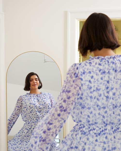 Plus size woman looking radiant in a mirror wearing a blue and white floral dress perfect for April Theory.