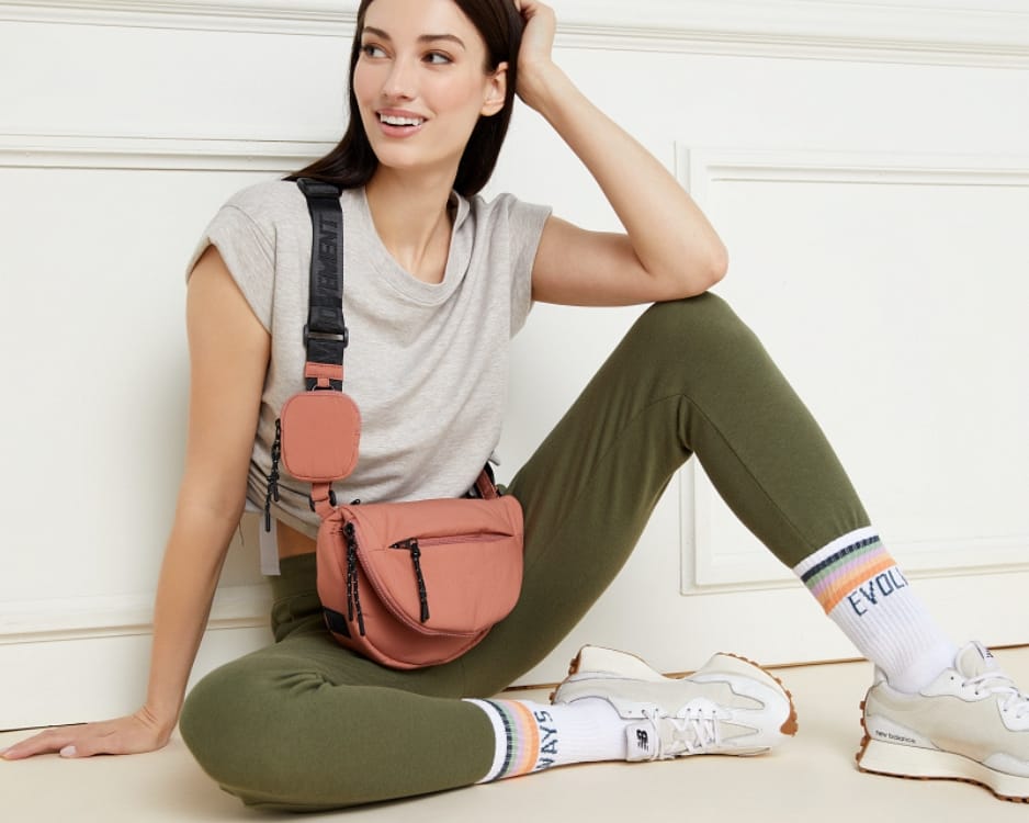 Brunette sitting against a wall wearing olive leggings, a grey tee, and a puffer sling bag.