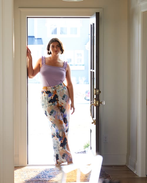Plus size woman standing in a doorway wearing a purple tank top and floral pants.