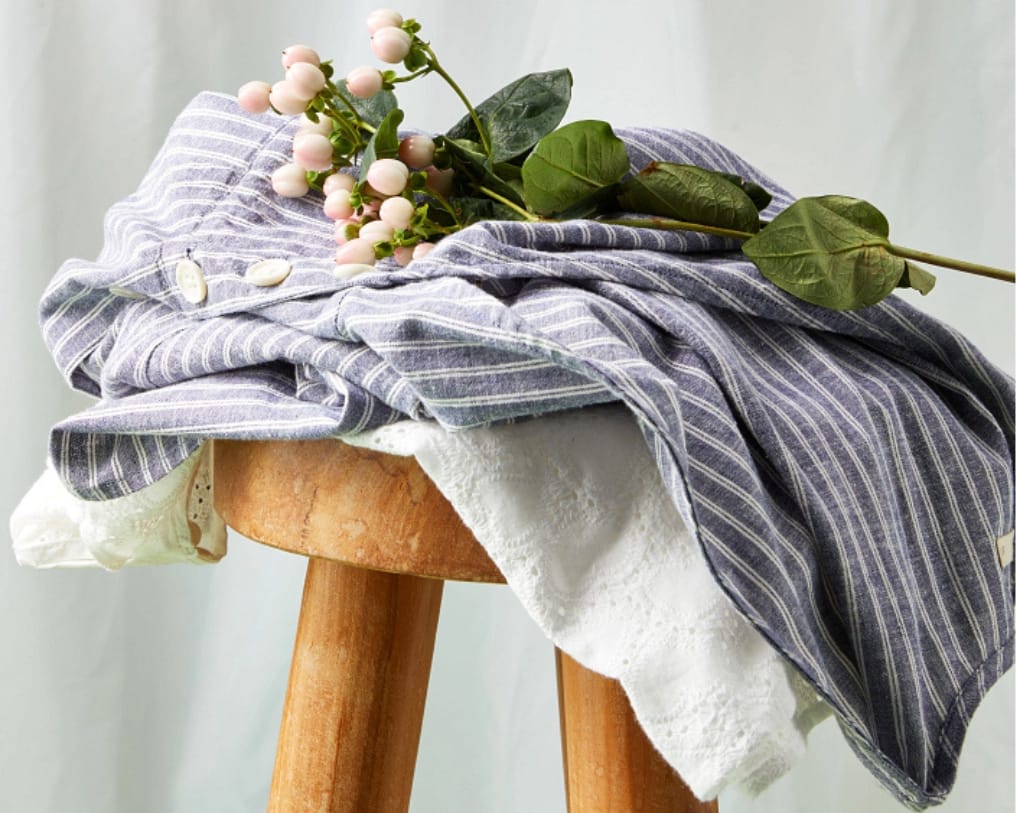 A white eyelet top and a blue and white striped dress perfect for dressing for April Theory folded and stacked on a wooden stool with a sprig of budding flowers sitting on top.