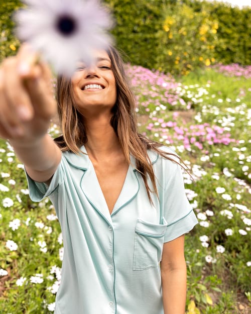 Smiling brunette in a flower field wearing green pajamas, holding a flower up to the camera.