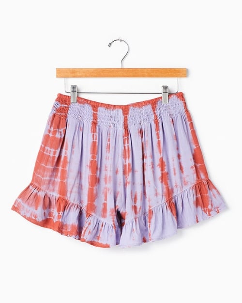 An up-close photo of the back of a pair of tie-dyed ruffle shorts. They are hanging on a wooden hanger in front of a white wall. 