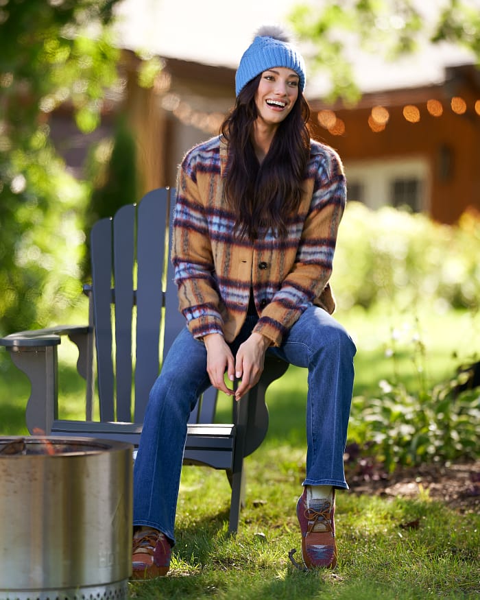 Laughing brunette sitting on patio furniture outside at a backyard bonfire party wearing a beanie, black jacket, and jeans.