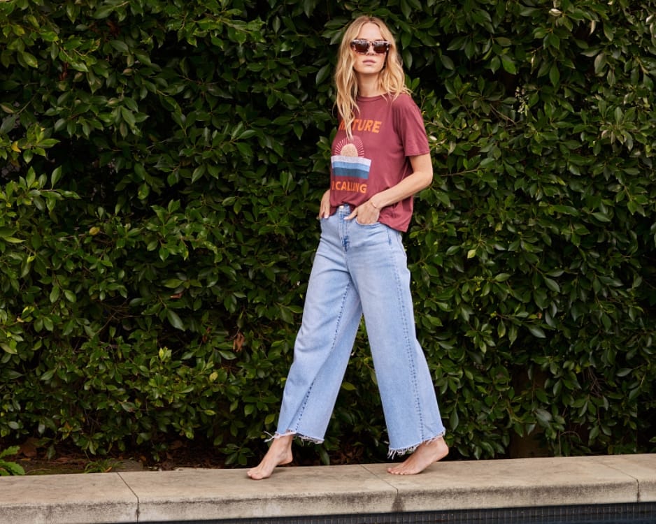 Blonde walking along a pool's edge at a backyard party wearing sunglasses, a graphic t-shirt, and light wash wide leg jeans.