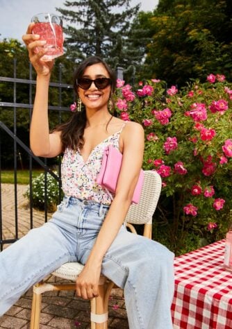 Brunette at a backyard party toasting the camera while wearing a floral blouse and light wash jeans.