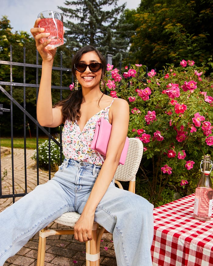 Smiling brunette wearing sunglasses, a floral blouse, and light wash wide leg jeans sitting outside at a picnic table, toasting at a backyard party.