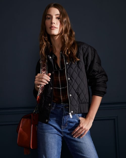 Model wearing a black quilted bomber with jeans and a suede brown bag with an embroidered strap. 