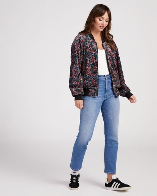 Model wearing a suede, floral patterned bomber jacket paired with jeans and black Adidas sneakers. 