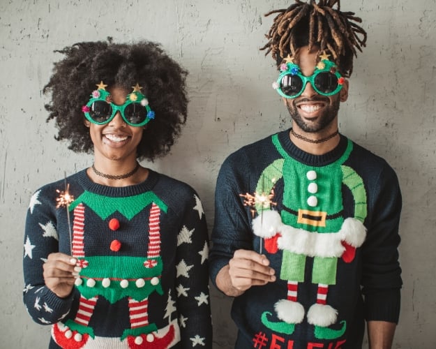 A man and a woman with dark brown hair and dreadlocks wearing ugly Christmas sweaters, Christmas tree sunglasses, and holding sparklers. 