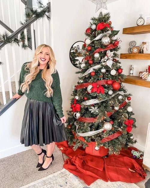 A girl with long blond hair and red lipstick standing near a Christmas tree dressed in a leather pleated midi skirt, a green sweater, and strappy black heels. This is how to dress for a Christmas party!