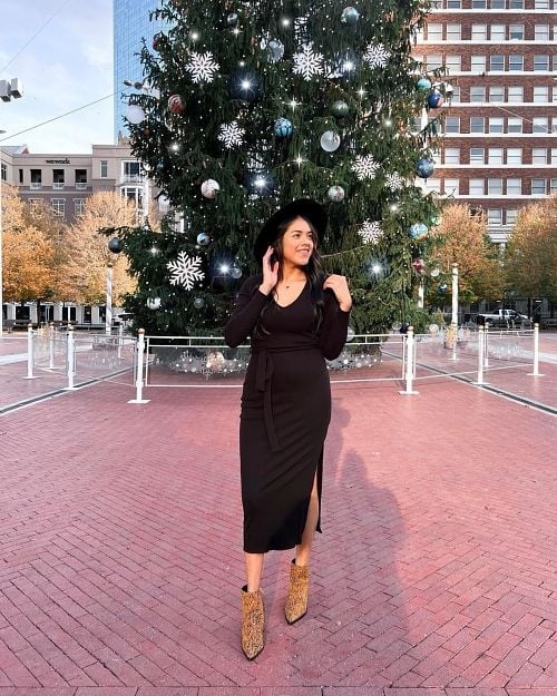 A girl with dark brown hair wearing a black midi dress, leopard-print ankle boots, and a black hat standing outside in front of a huge Christmas tree.