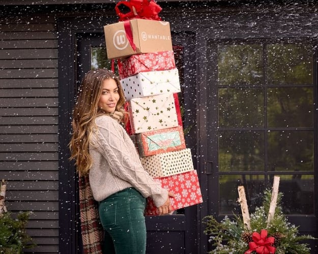 A model with long brown hair dressed in a chunky oatmeal sweater and emerald green corduroys holding a stack of Christmas presents in the snow.