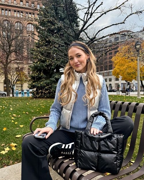 A girl with long blond hair in a headband sitting on a park bench wearing a blue sweater, a silver puffer vest, black pants and black sneakers.