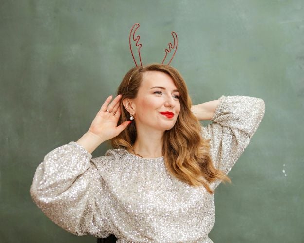 A woman in red lipstick and a festive antler headband wearing a silver sequined long-sleeved top. 