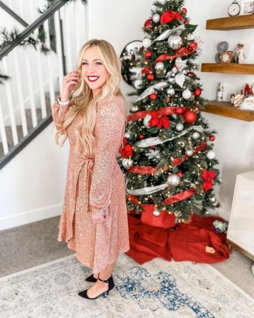 An influencer with long blond hair standing in front of a Christmas tree wearing a sequined pink wrap dress and strappy black heels.